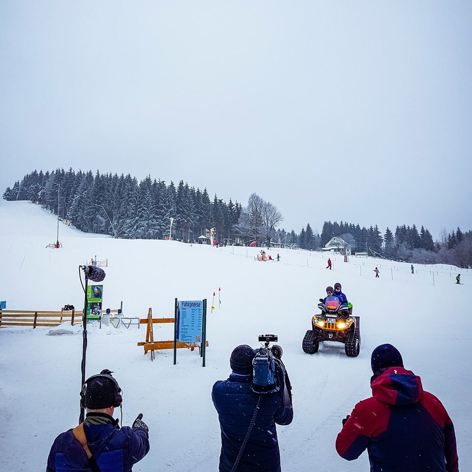 Zwei Männer der Bergwacht fahren mit dem Kettenquad über den Schneehügel. Das Kamerateam steht am Bildrand und filmt.
