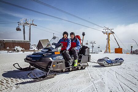 Zwei Bergretter sitzen auf einem Schneefahrzeug neben einem Skilift. Sie sichern die Skipiste ab.