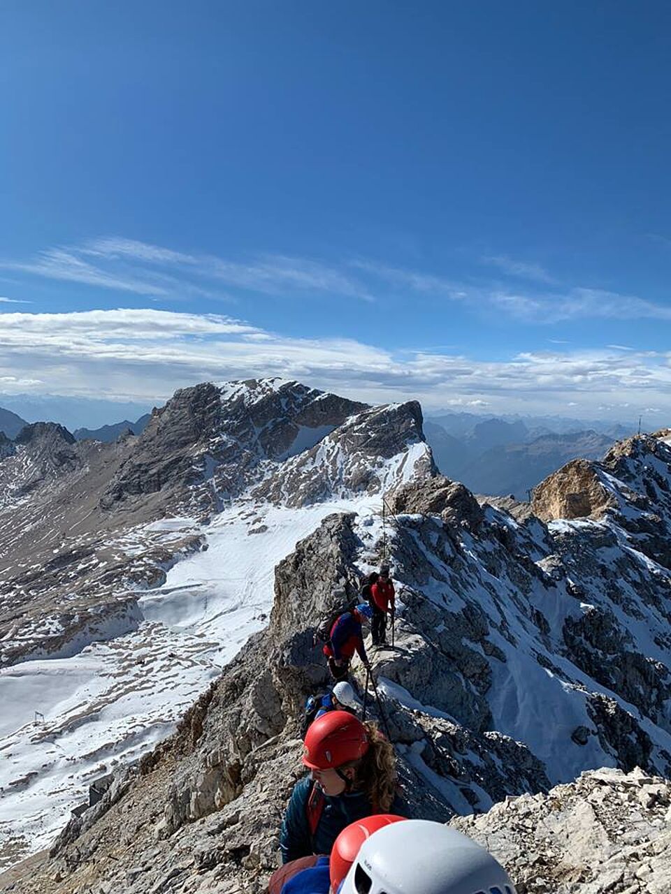 Mehrere Personen erklimmen eine schneebedeckte Bergspitze. Sie wandern entlang eines Sicherungsseils bei strahlend blauem Himmel.