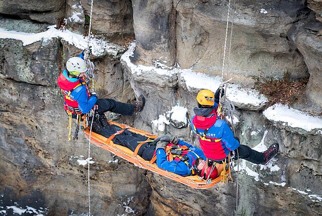 Zwei Bergwachtretter transportieren eine verletzte Person in einer Trage am Hang eines Felsens.
