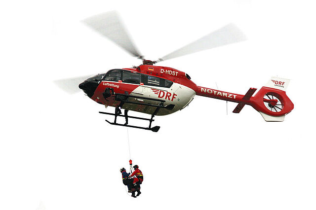 Ein Hubschrauber steht in der Luft. Zwei Bergretter hängen an einem Seil unter ihm.