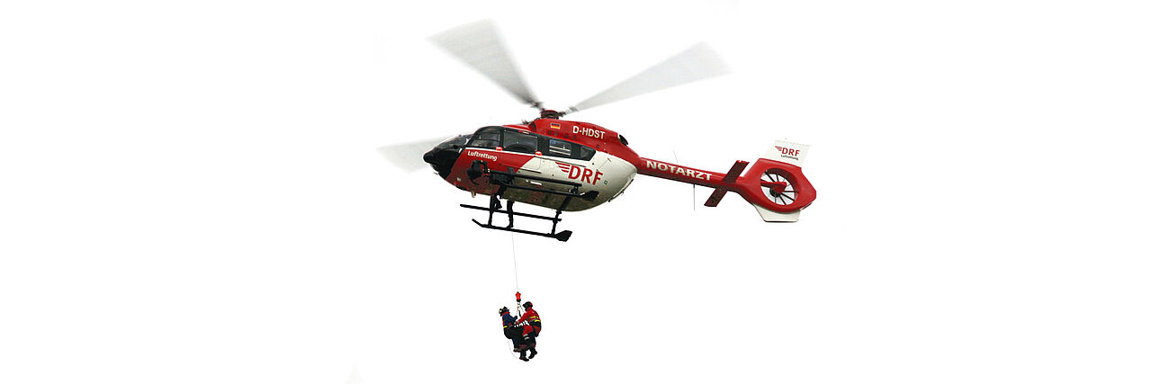 Ein Hubschrauber steht in der Luft. Zwei Bergretter hängen an einem Seil unter ihm.